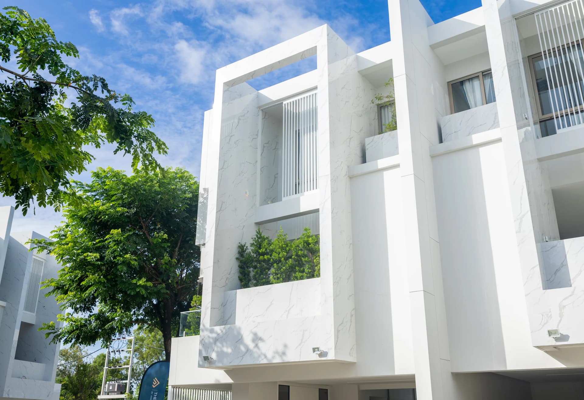 Eine fertige Villa: weiße Fassade in Marmoroptik, der begrünte Balkon im 1. Obergeschoss und alte Bäume davor